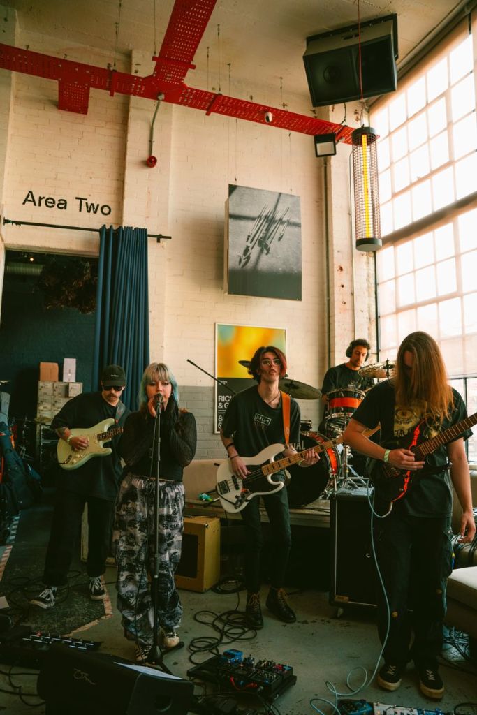 A band performing live on stage in a modern venue, with a female vocalist holding a microphone, surrounded by guitarists and a drummer. The setting features industrial decor and large windows, with a sign reading 'Area Two' visible in the background.