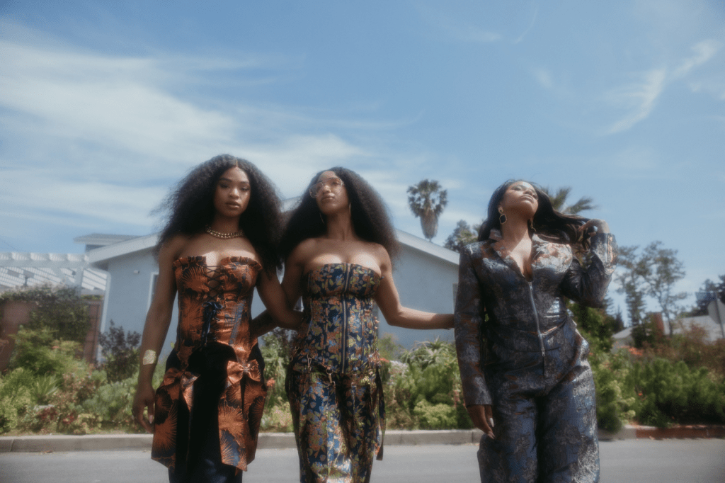Three women walking together in stylish outfits against a backdrop of greenery and a blue sky.