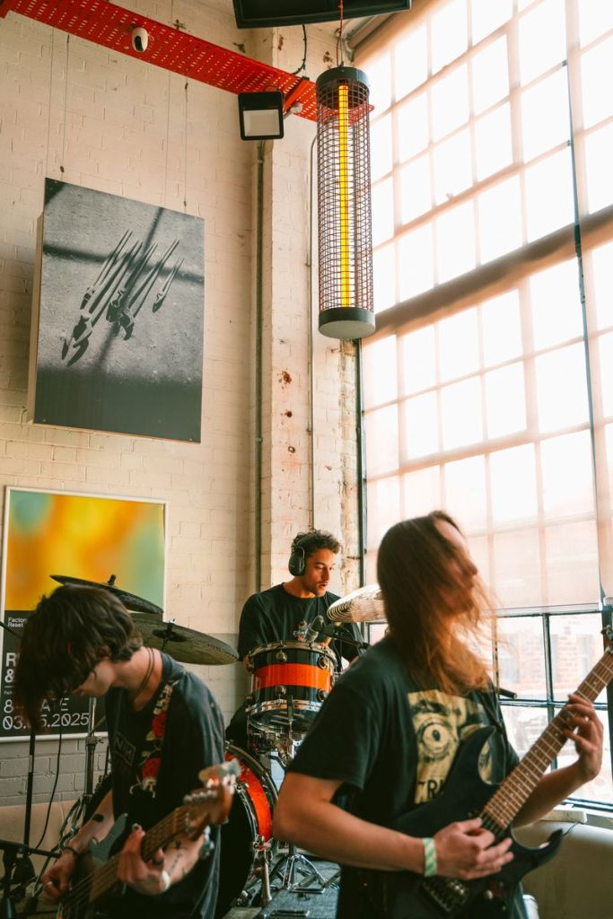 A band performing live in a well-lit venue, featuring a drummer in headphones, a guitarist playing an electric guitar, and a bassist. Modern decor includes art on the brick walls and large windows allowing natural light.